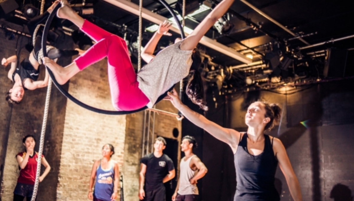 four people watch while two others hold themselves up on an aerial hoop and rope. A person helps them to balance.